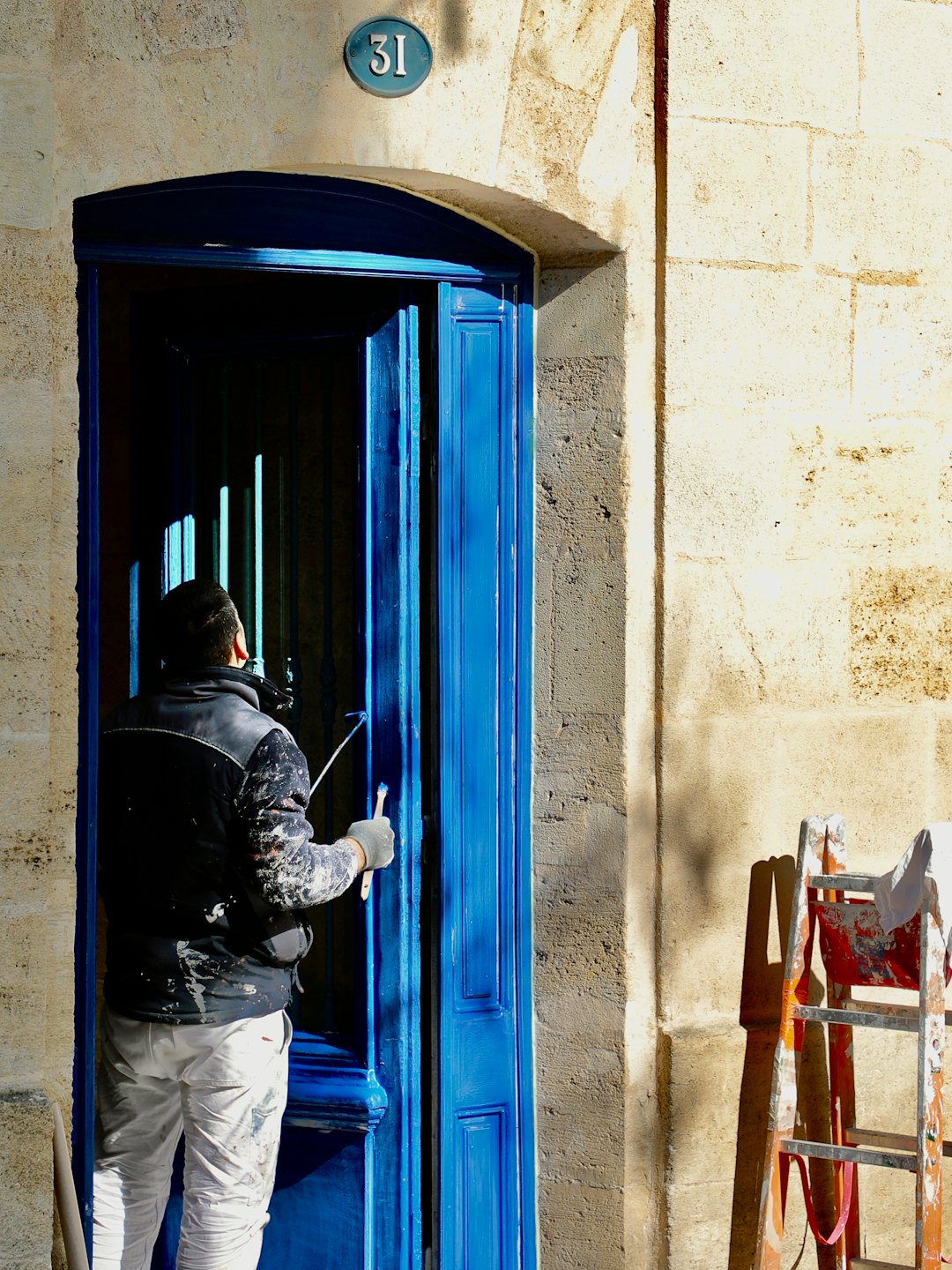 Een schilder werkt aan een blauwe deur van een stenen gebouw.